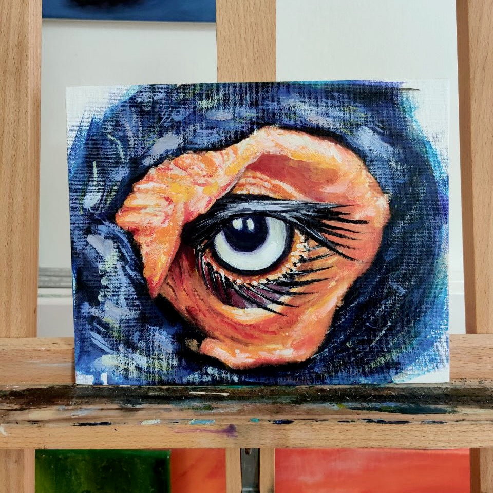 A piece of abstract art depicting an eye with predominantly blue and orange colors on a small oil board, displayed on a wooden easel.