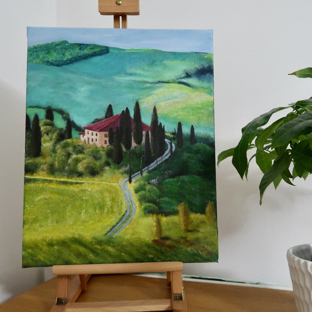 An oil on canvas painting depicting a landscape with rolling hills, greenery, and a rustic house, displayed on a wooden easel.