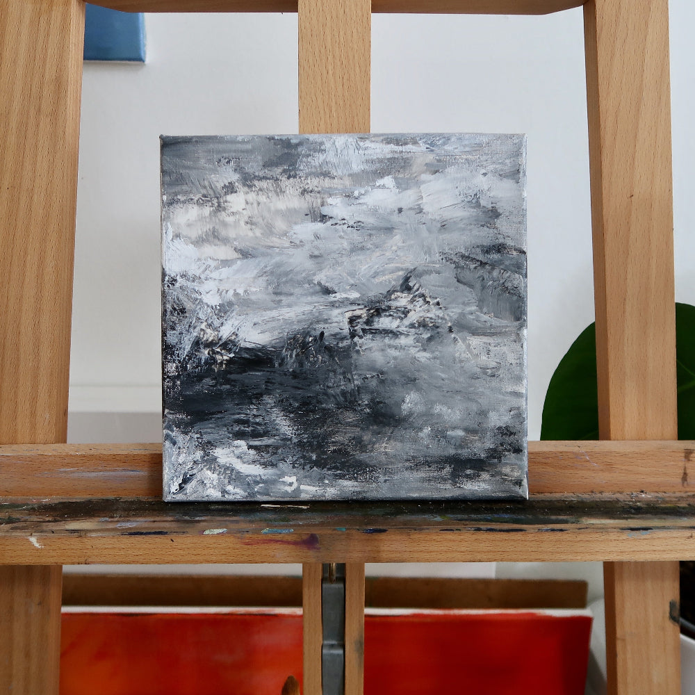 Abstract acrylic landscape painting displayed on a wooden easel, predominantly in shades of gray with a few splashes of other colors.