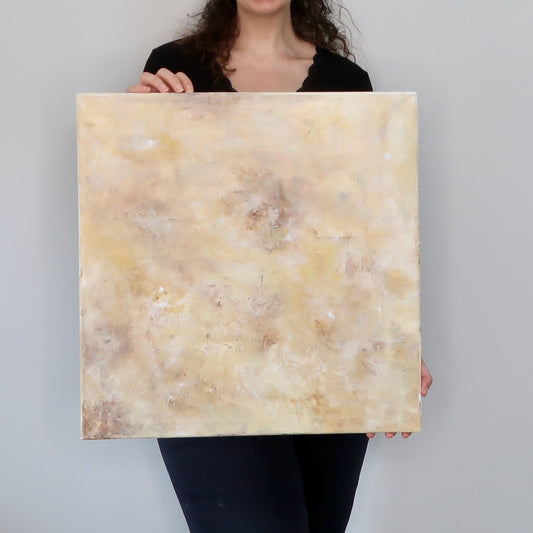A person holding a square abstract acrylic landscape painting in warm yellowish and earthy colors.