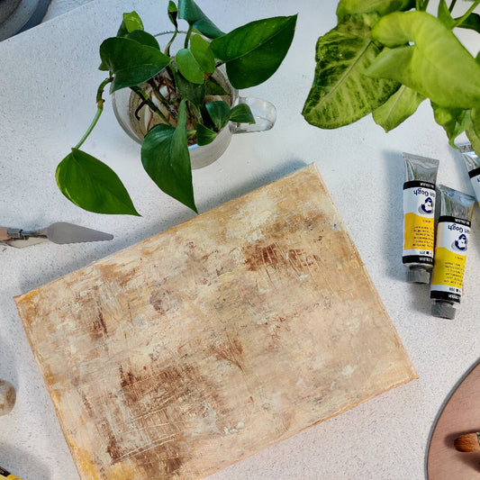 An abstract acrylic landscape painting laid out on a table, surrounded by various objects including a plant pot, with the artist's materials like brushes and paint tubes visible.
