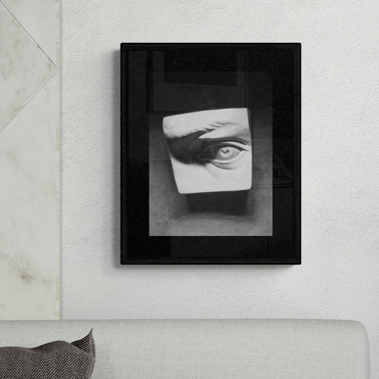 A charcoal drawing of an eye cast, displayed in a black frame, hung on a wall above a white sofa.